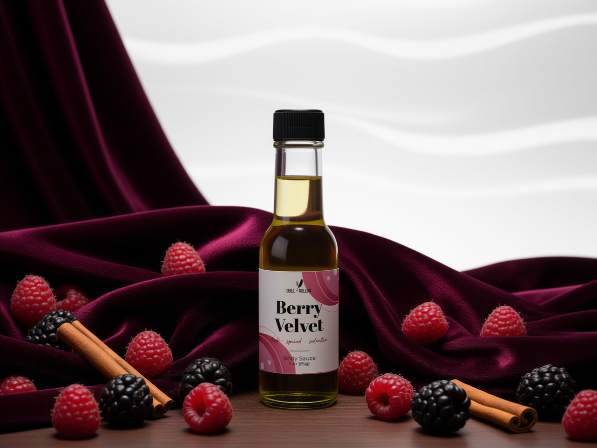 A bottle of Berry Velvet body oil stands centered on a wooden surface, surrounded by fresh raspberries, blackberries, and cinnamon sticks, with draped deep burgundy velvet fabric and a white background.