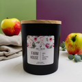 Black candle with a wooden lid and floral label, surrounded by apples and eucalyptus leaves on a white surface.