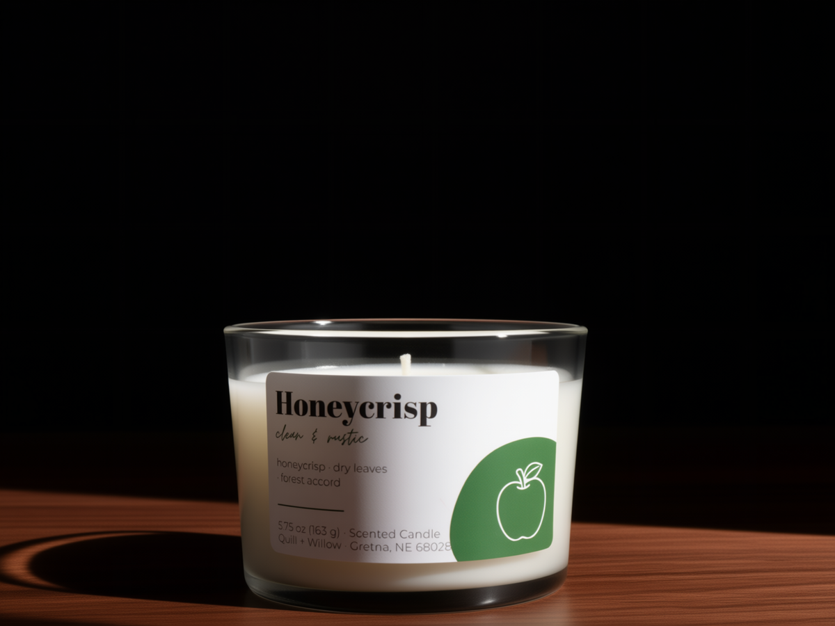 A glass jar candle labeled 'Honeycrisp' with white wax and a green apple graphic, set on a wooden surface against a black background.