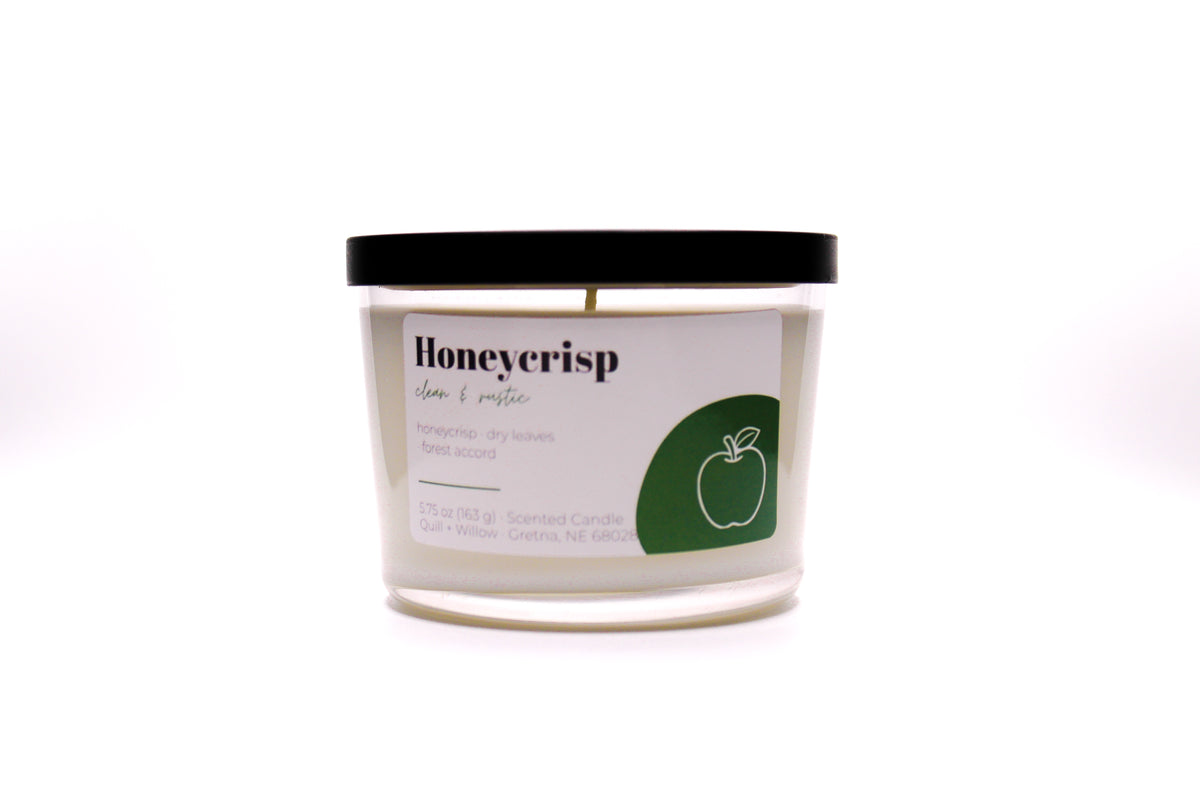 A scented candle in a clear glass jar with a black lid and a white label featuring a green apple graphic. The label reads 'Honeycrisp' and 'Apple + pine' with 'Scented Candle' below, set against a plain white background.
