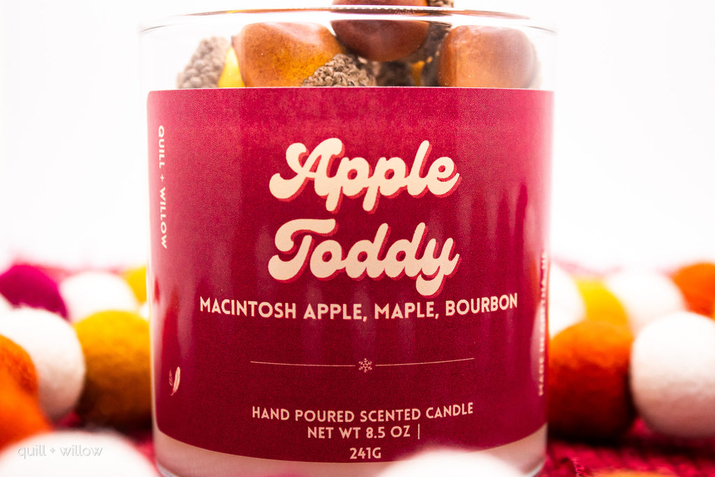 Candle jar labeled 'Apple Toddy' with colorful elements in the background