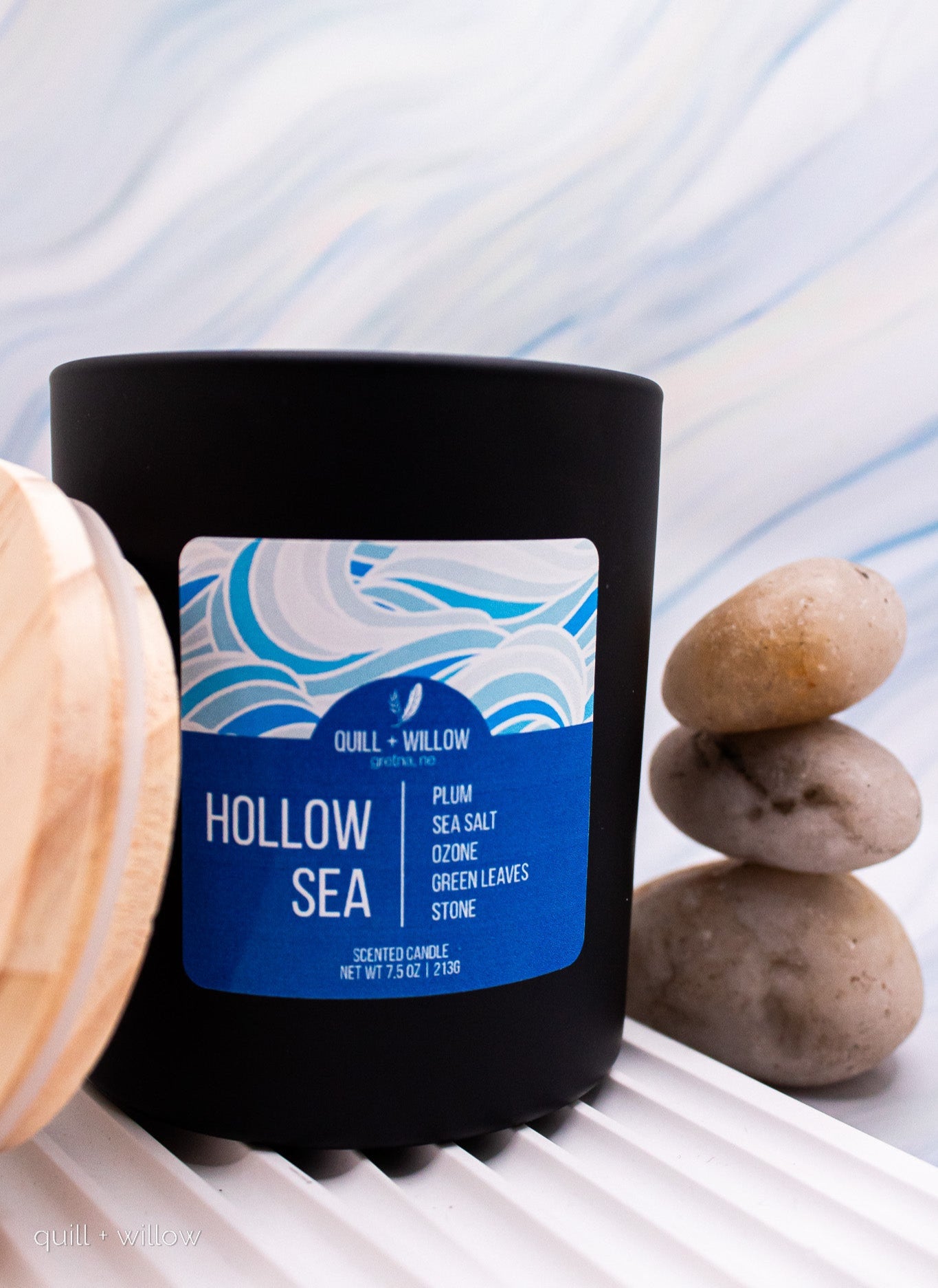 Black candle with a blue label titled 'Hollow Sea' on a white surface with stones.
