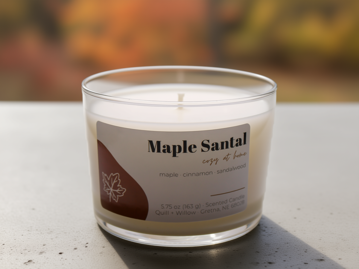 A single-wick scented candle in a clear glass jar labeled 'Maple Santal cozy home' on a light stone surface with a blurred outdoor autumn background.