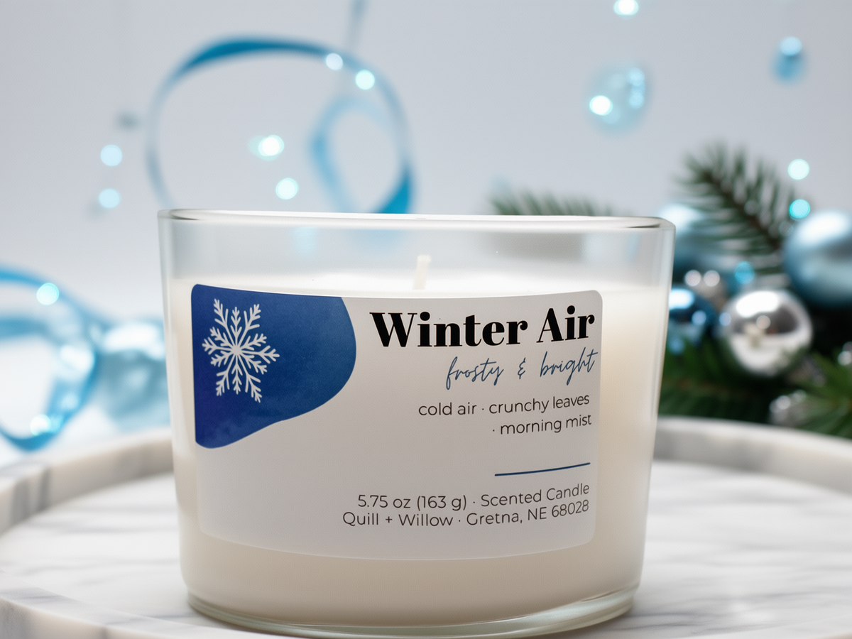 A clear glass jar candle labeled 'Winter Air' sits on a white marble surface with a white and blue label. The candle is photographed in a styled holiday setting with blurred blue ornaments and greenery in the background.