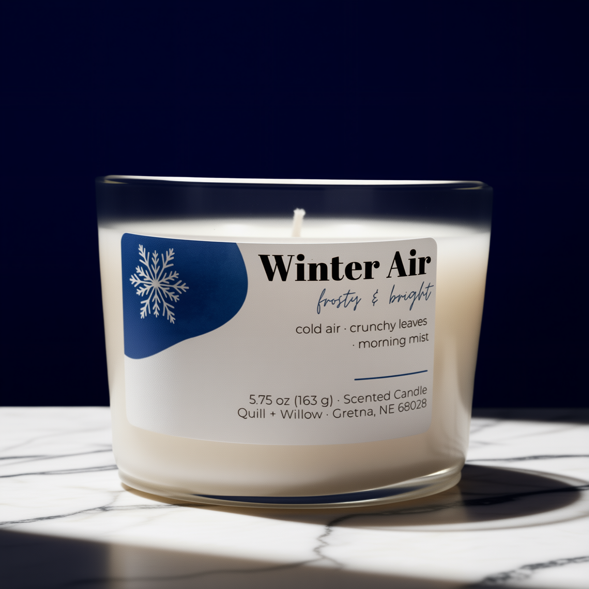 A scented candle in a clear glass jar with a white wax fill and a white label featuring a blue snowflake graphic and the text 'Winter Air frosty & bright.' The candle sits on a white marble surface against a dark navy background.