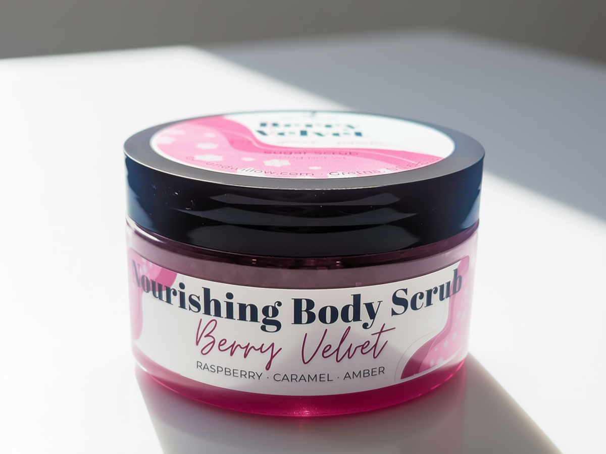 A jar of Nourishing Body Scrub in Berry Velvet scent, featuring a translucent pink container, a black lid, and a white label with pink accents, set on a white surface under direct sunlight.